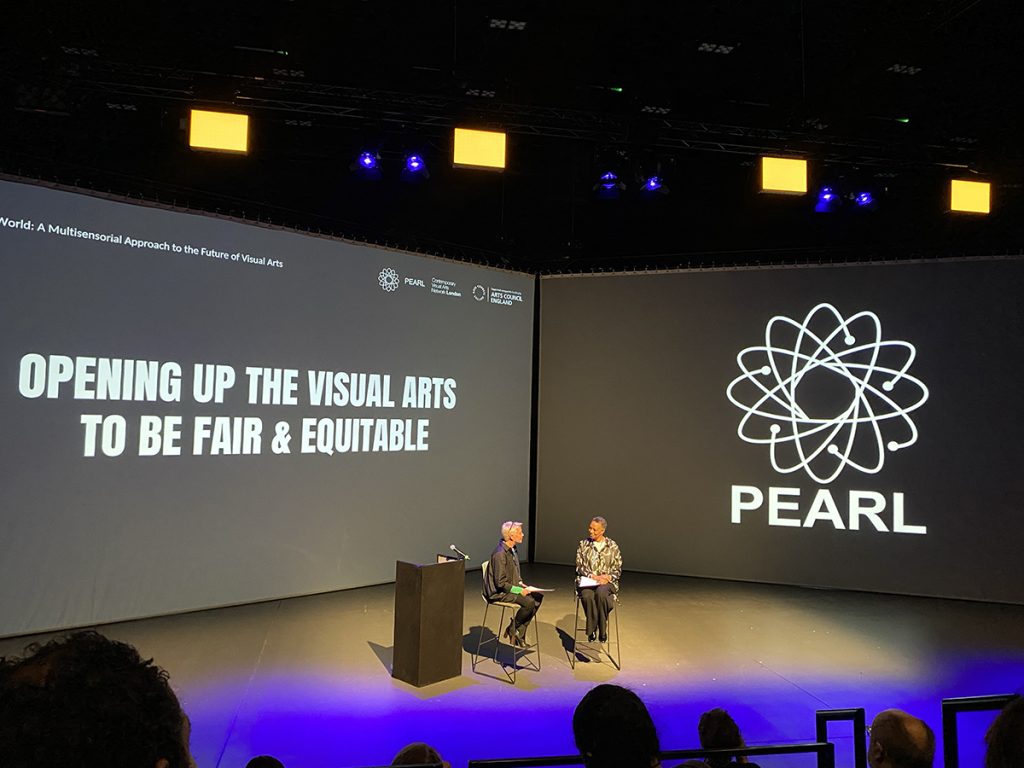 Sheryll Catto sits on a chair opposite a woman who is talking to her. They are in front of an audience, and there is projected text in the background that says 'opening up the visual arts to be fair and equitable'.