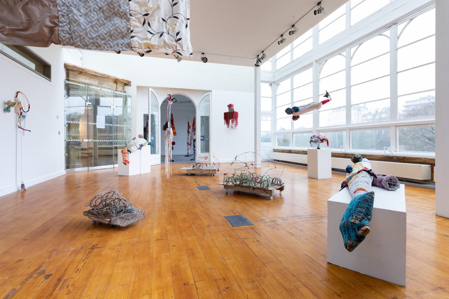 An exhibition space with multiple sculptures, some suspended from the ceiling, on plinths and the floor.
