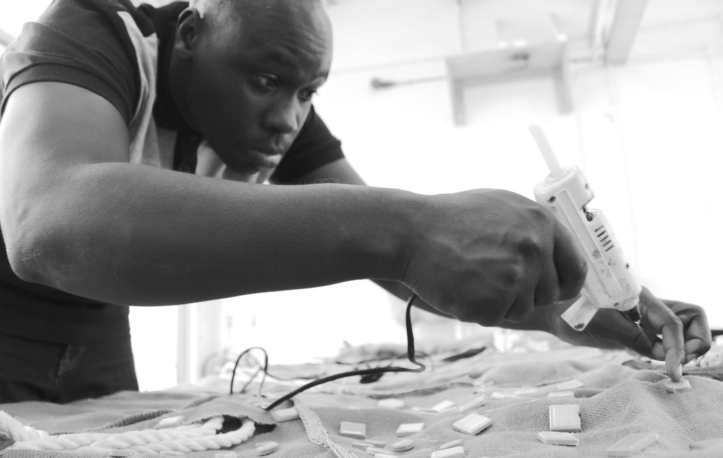 A black and white photograph of artist Andrew Omoding sticking mosaic tiles to a piece of fabric using a glue gun.