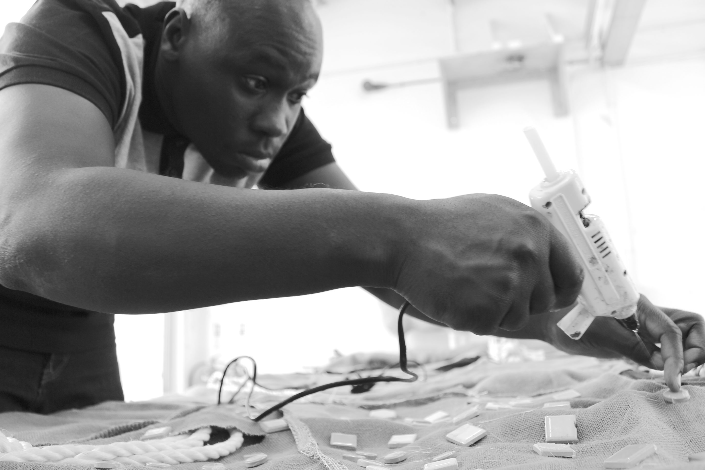 A black and white photograph of artist Andrew Omoding sticking mosaic tiles to a piece of fabric using a glue gun.