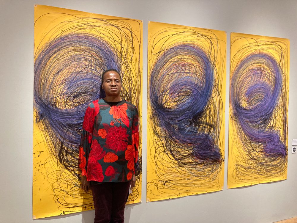 Nnena Kalu standing in front of three of her artworks next to each other on a wall, showing repeated swirling marks on yellow paper.