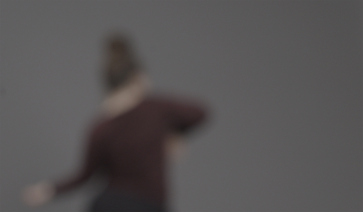 A blurred image of a figure. They are wearing a dark top and they have their back to the camera, and the background around them is dark grey.