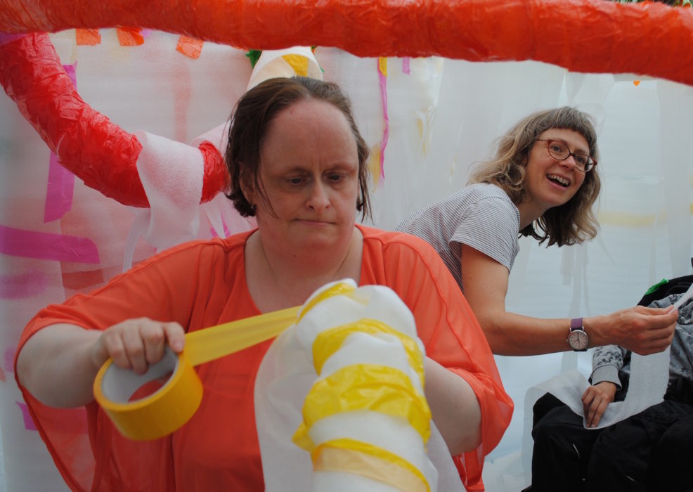 Linda Bell tapes her artwork together. A volunteer smiles in the background.