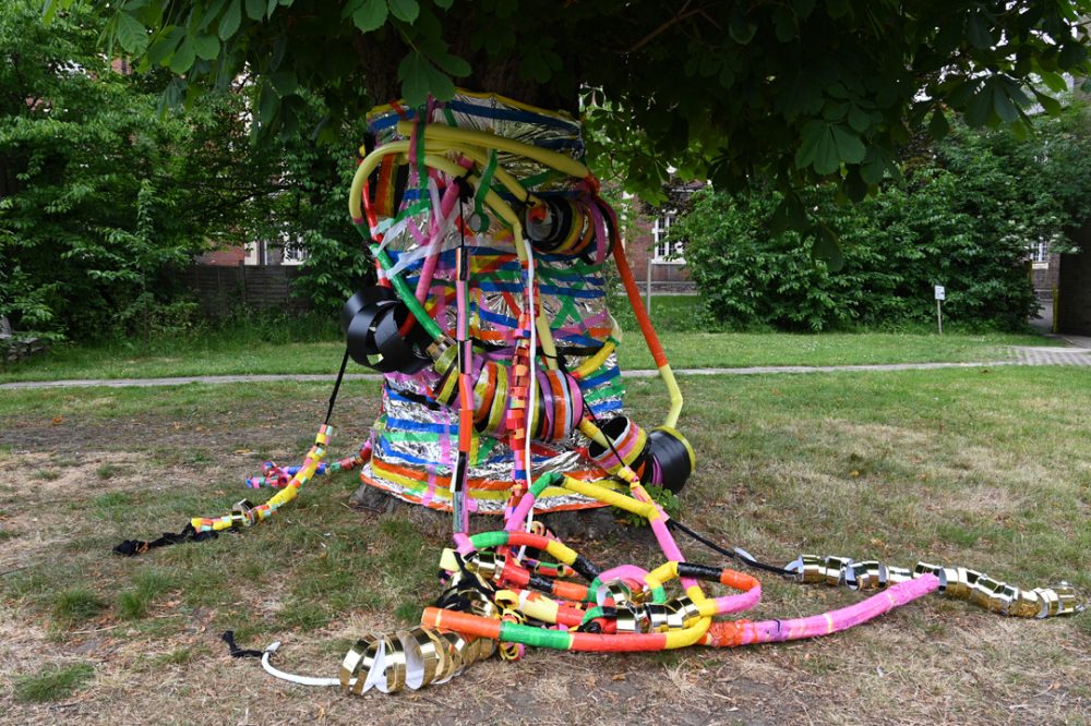 Linda Bell's large sculptural piece, displayed outside in a park. The artwork is a range of colourful tubes, with colourful tape wound around it.