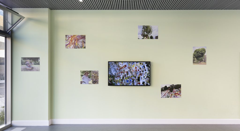 Linda Bell's work in a gallery space. The image shows one painting in the middle of the wall, with photos of Linda Bell around it.