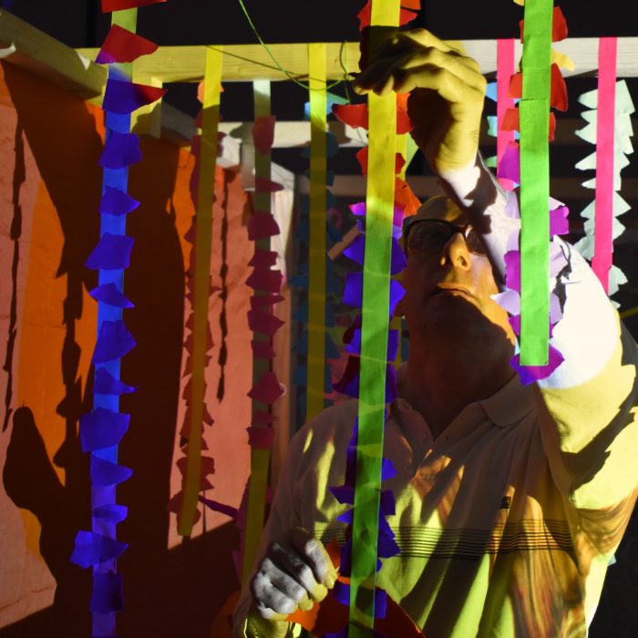 Mark Lawrence reaches upwards to hang a strip of fabric to a wooden frame. The light of a projector makes the room yellow and red coloured.