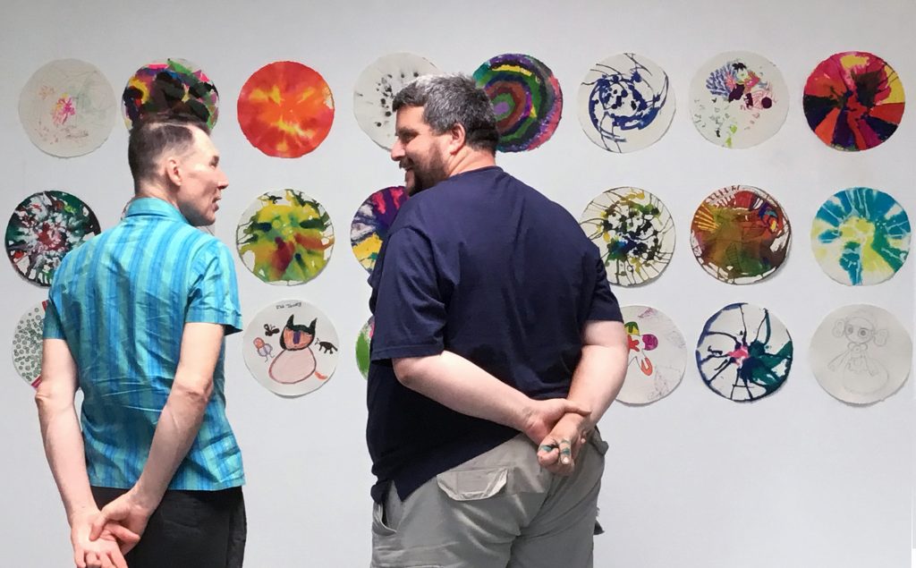 Two men stand in front of a wall, talking to each other. Behind them are a selection of ink and pencil drawings on small circular pieces of paper.