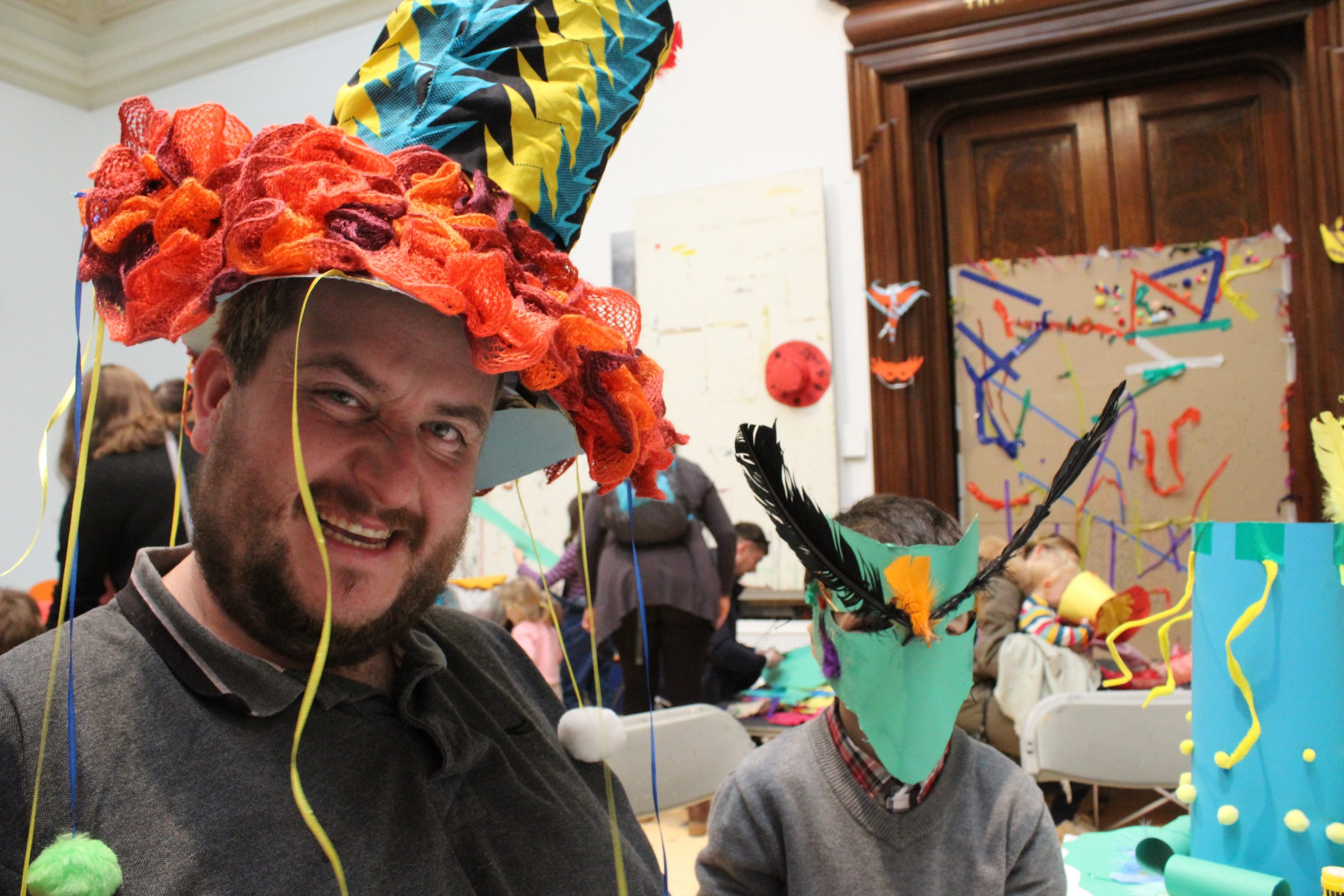 Artist Matthew Berry wearing a custom made hat stood next to a child with a DIY paper mask at one of his workshops.