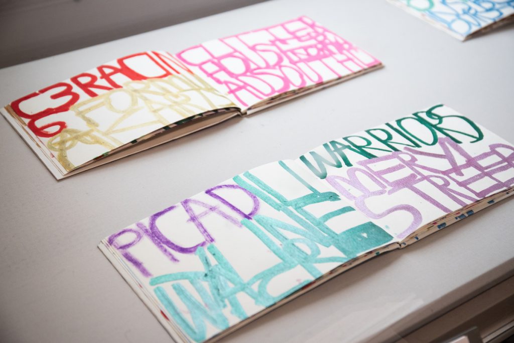 2 sketchbooks lay open on a table, with large colourful writing in them, with words such as 'warriors' and 'meryl streep'.
