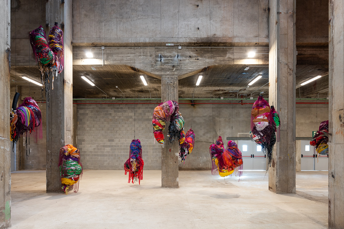 Eight brightly coloured sculptures made of wrapped strips of material hang at differing heights in a brutalist exhibition space.