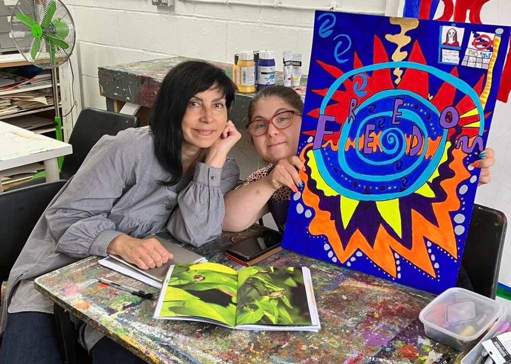 Ophir Yaron sits next to Jo Longhurtst. She is in the studio sat behind a table, and is holding up one of her artworks and smiling.
