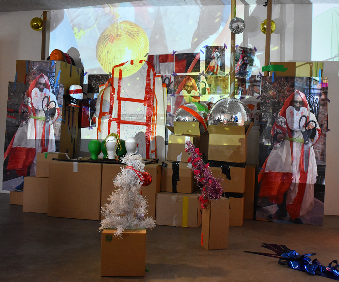 Pardip Kapil's exhibition. There is a range of objects, including brown cardboard boxes, a small white christmas tree, a disco ball and some mannequin heads. On the wall behind is a projected image of a gold disco ball.