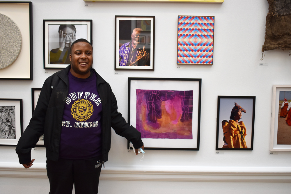 Patrick stands near his framed artworks, smiling at the camera.
