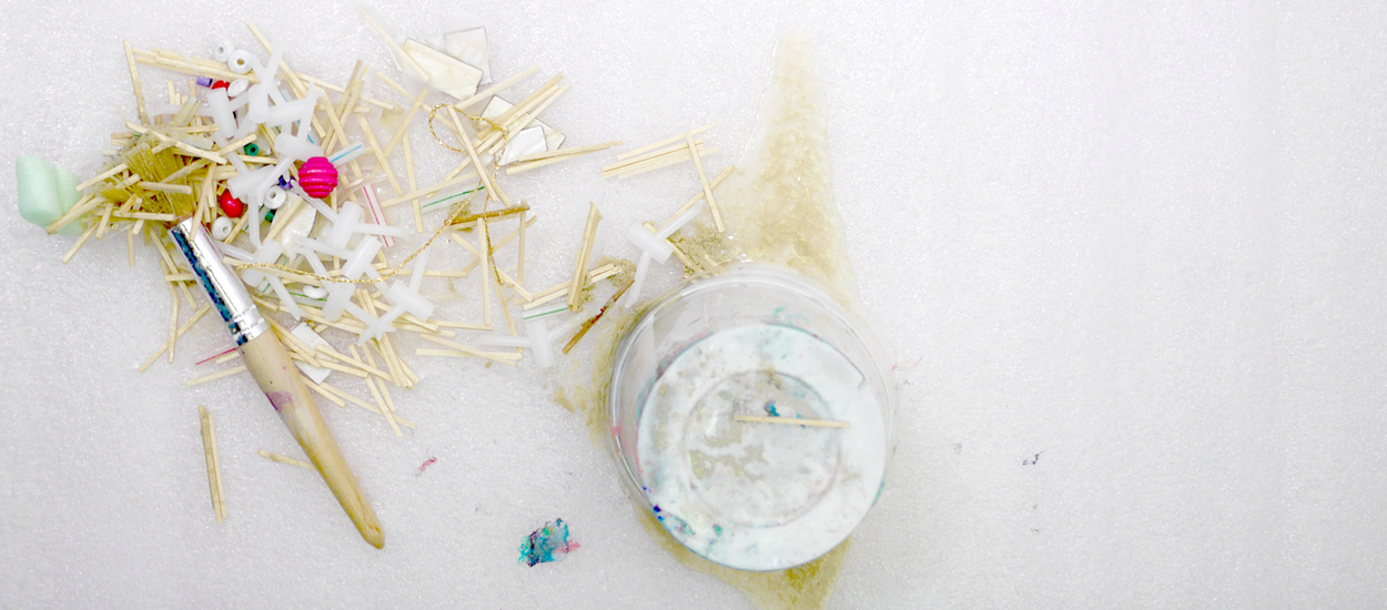 A brush and glue pot are photographed on a white background. A heap of wooden sticks and white plastic objects are placed on top.