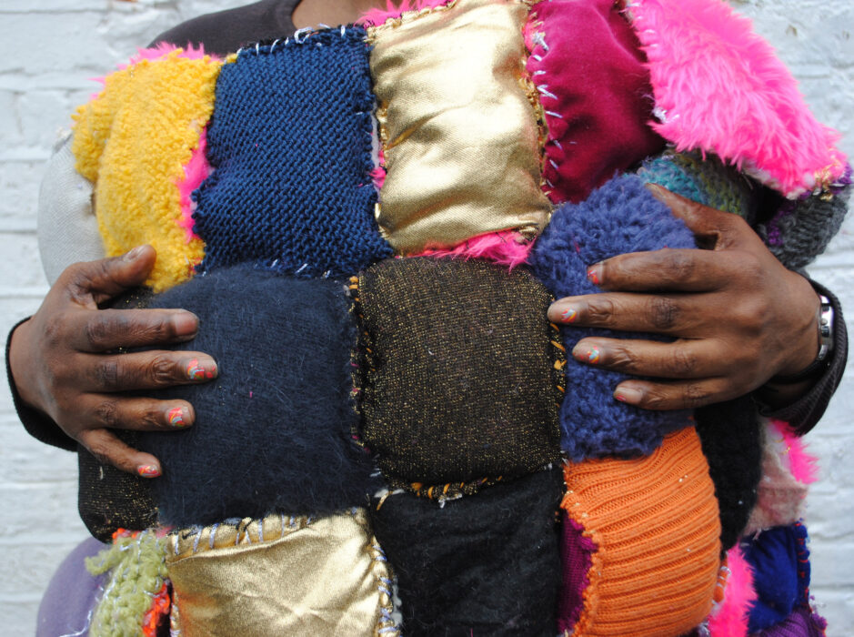 Lasmin holds her artwork to her body. We can see her hands holding onto the colourful patchwork quilt.