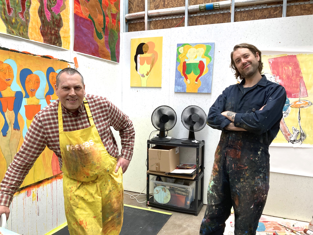 Leaning on a table, artist Robin Smith stands in his studio space wearing a yellow apron. Beside him is artist Richard Phoenix.