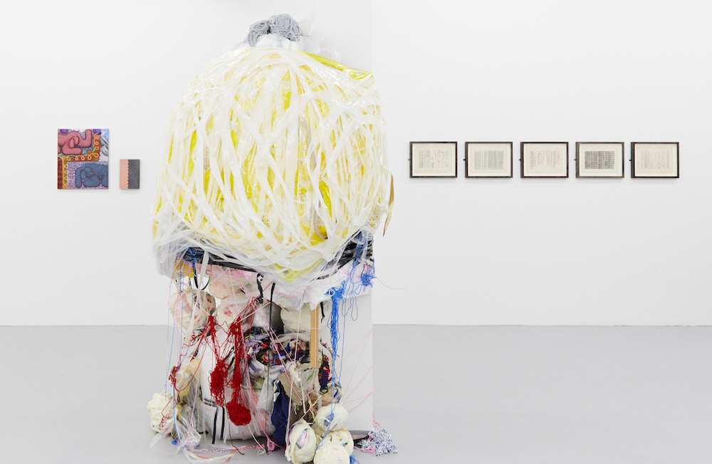 A large, multimedia sculpture made of bundled strips of material and yarn sits in a white gallery space in front of wall mounted works.