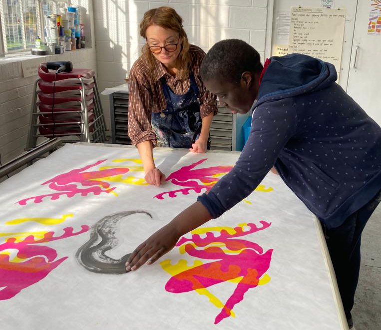 Sharon Adokorach works with colleague on an artwork. It's a large piece of white fabric on a table with pink and yellow printed shapes on it. Sharon is reaching to the middle of the fabric, where there is a grey pattern.