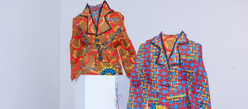 Two colourful, textural suit jackets are displayed in a gallery setting. One is orange, with painted red rings and black buttons, and the other is blue, with red and yellow chains painted on.