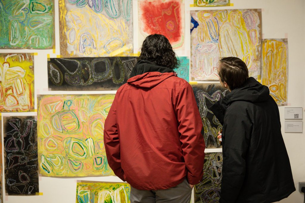 Two people stand looking at the artworks on the wall in a gallery space. The art is a selection of unframed, colourful drawings.
