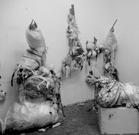A black and white photo of an art installation featuring sculptures bound in plastic wrap and tape. A large one leans against the wall next to a smaller one mounted against the wall. Another sits on a low plinth in the foreground.