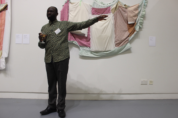 Artist Andrew Omoding stood in front of a textile artwork hung up on a wall, with his right arm pointing outwards and a drink in his left.
