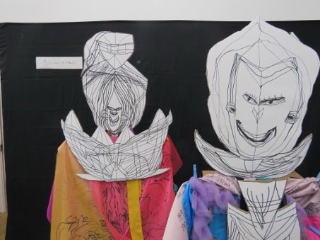 Two people wearing paper masks with black line drawing, whilst draped in colourful fabrics that have pens marks on.