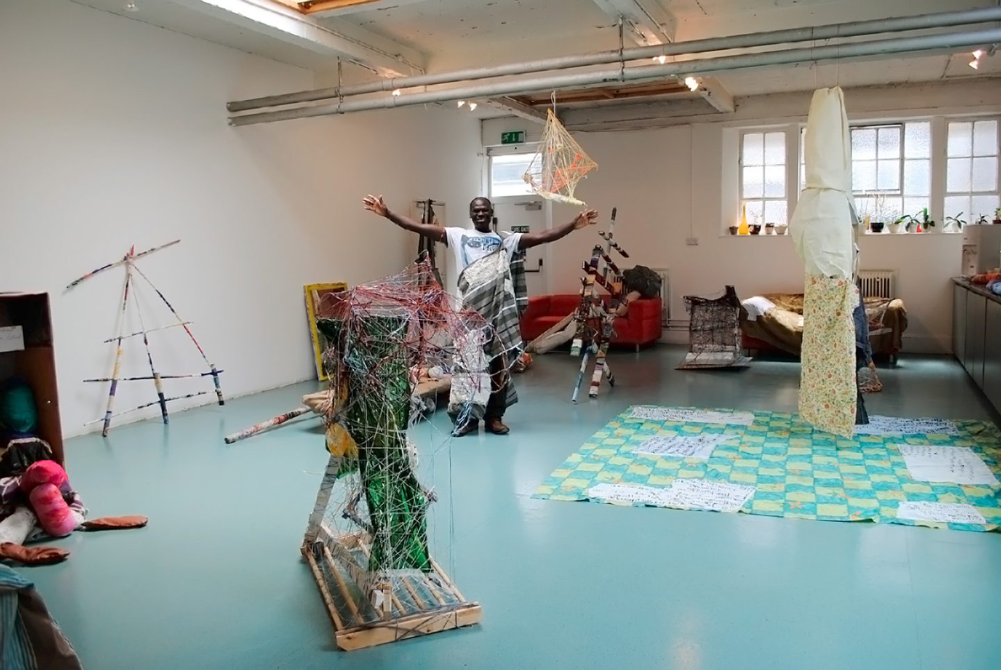 Artist Andrew Omoding posing with his arms stretched open in a studio space surrounded by his sculptures.