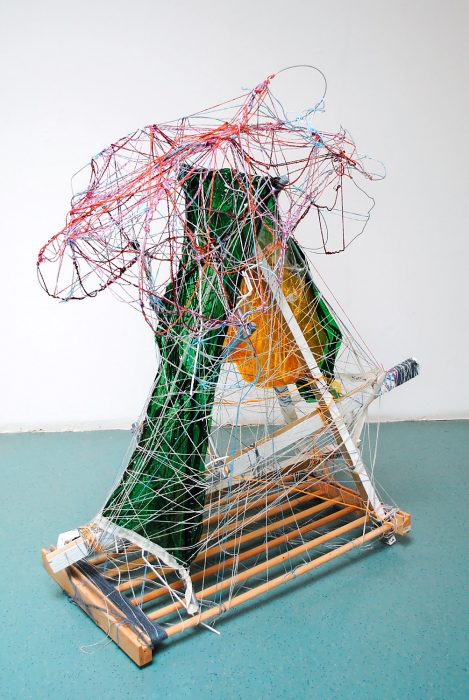 A sculptural work of Andrew's is shown. Wooden sticks are bound together with dark blue string, and two green pieces of fabric are sewn together on top to create a triangular shape. Pink, red and blue string is wound round the entire piece.