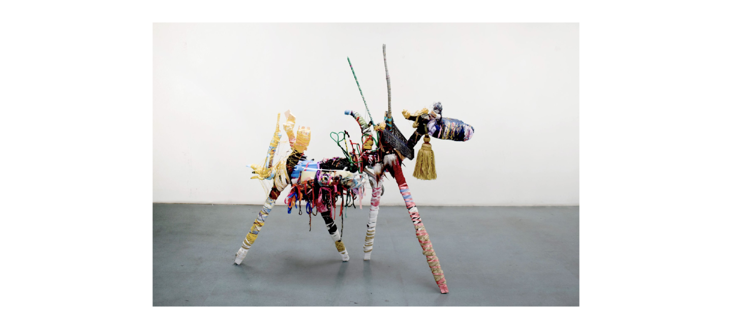 A mixed media sculpture stood on a grey concrete floor in a white walled room. The sculpture, resembling a goat, is made of various materials such as pink ribbon, gold tassel, blue foam etc.