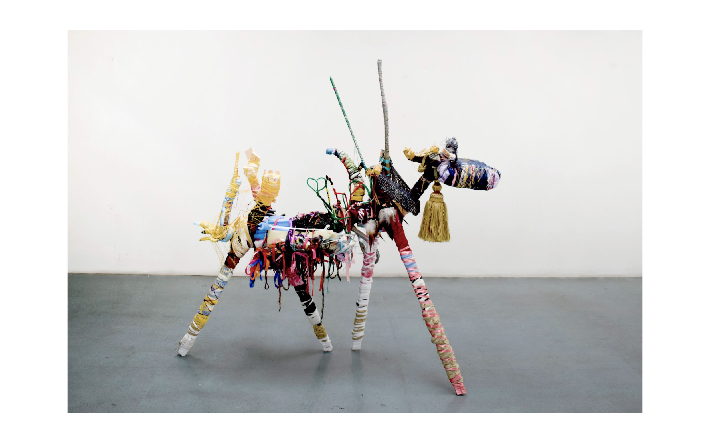 A mixed media sculpture stood on a grey concrete floor in a white walled room. The sculpture, resembling a goat, is made of various materials such as pink ribbon, gold tassel, blue foam etc.