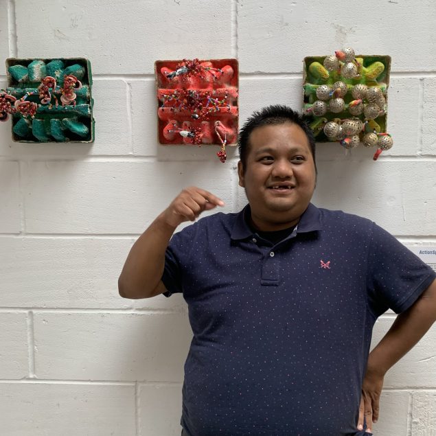 Abdul is wearing a blue t shirt and denim shorts, and is standing in front of several of his paintings and artworks mounted on a white wall.
