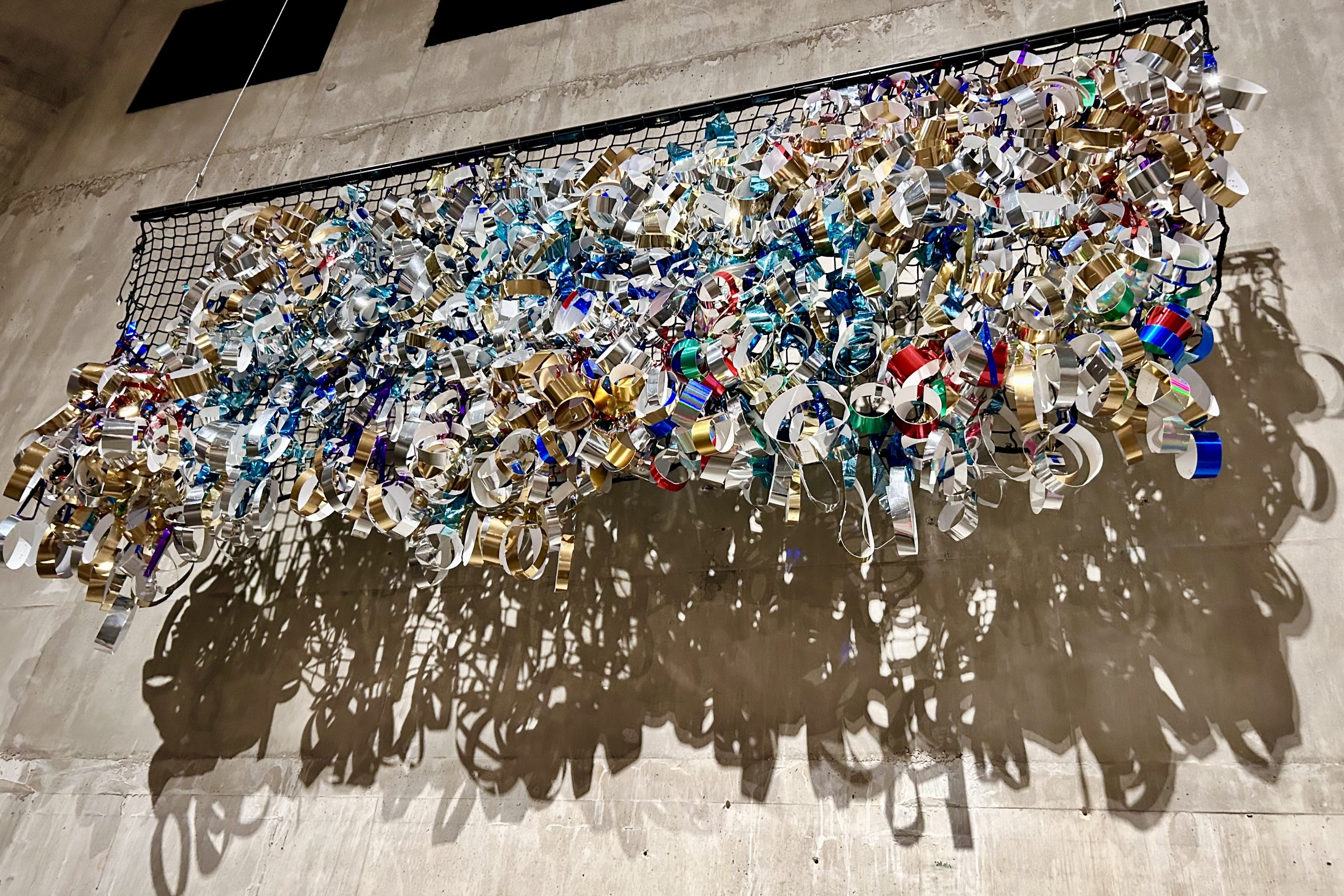 many rolls created from foil tape are mounted onto a black net, the net is suspended from the ceiling against the concrete walll of a tank in Tate Modern.