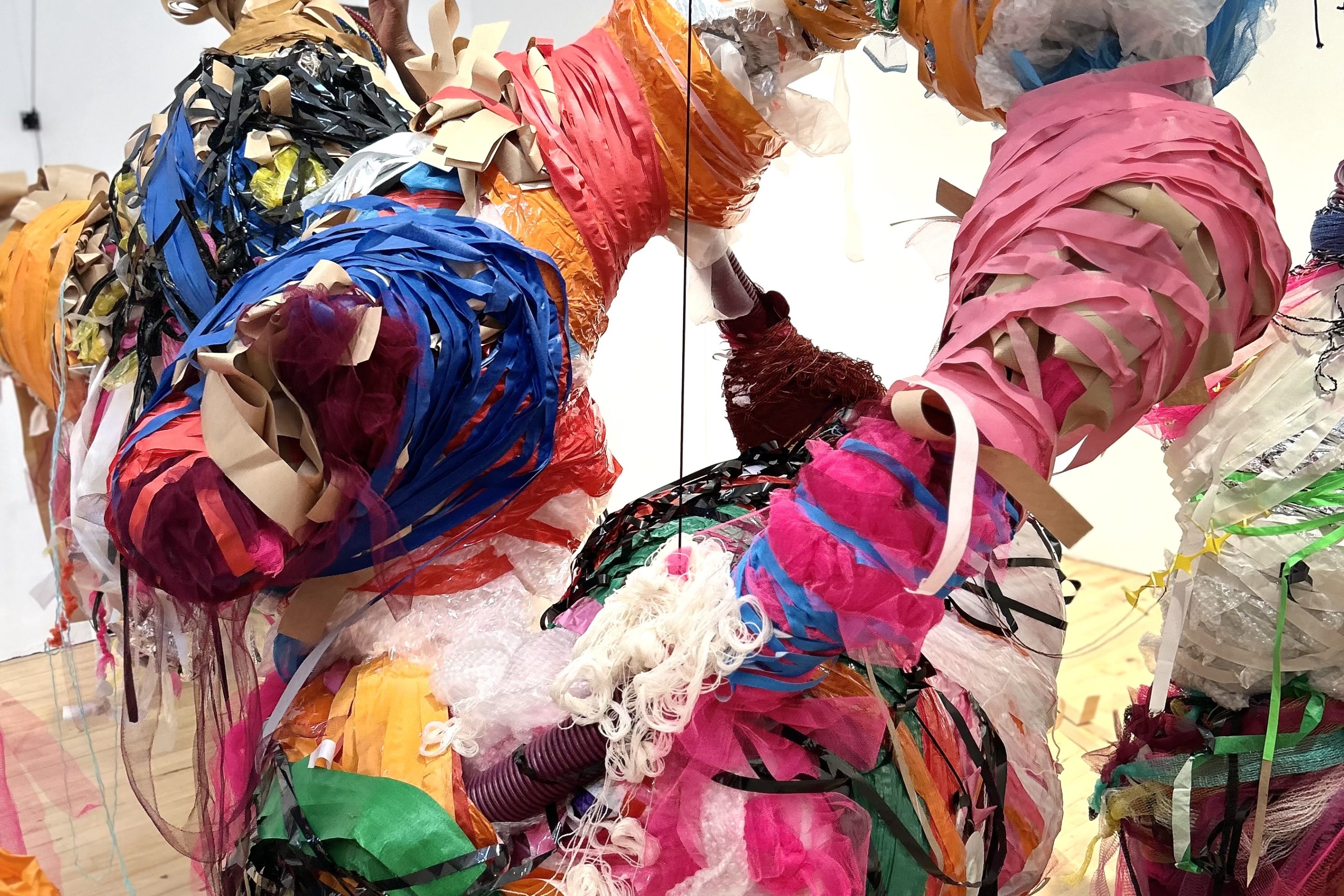 A densely wrapped hanging sculpture in bold colours suspended by wires from the ceiling.