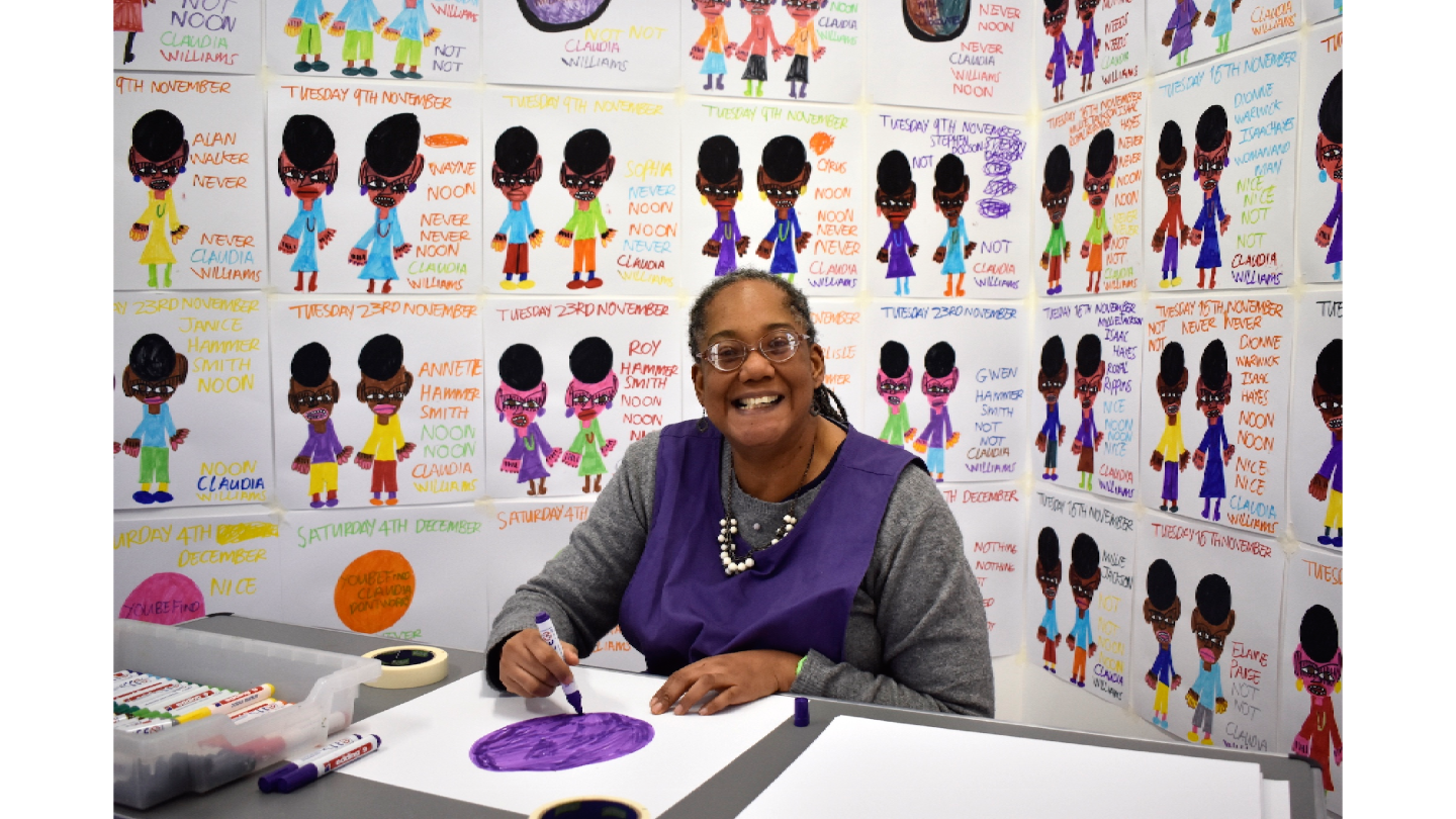 Photograph of Claudia sat at a table drawing. Claudia is looking at the camera smiling. She is surrounded by her drawings which are up on the wall.