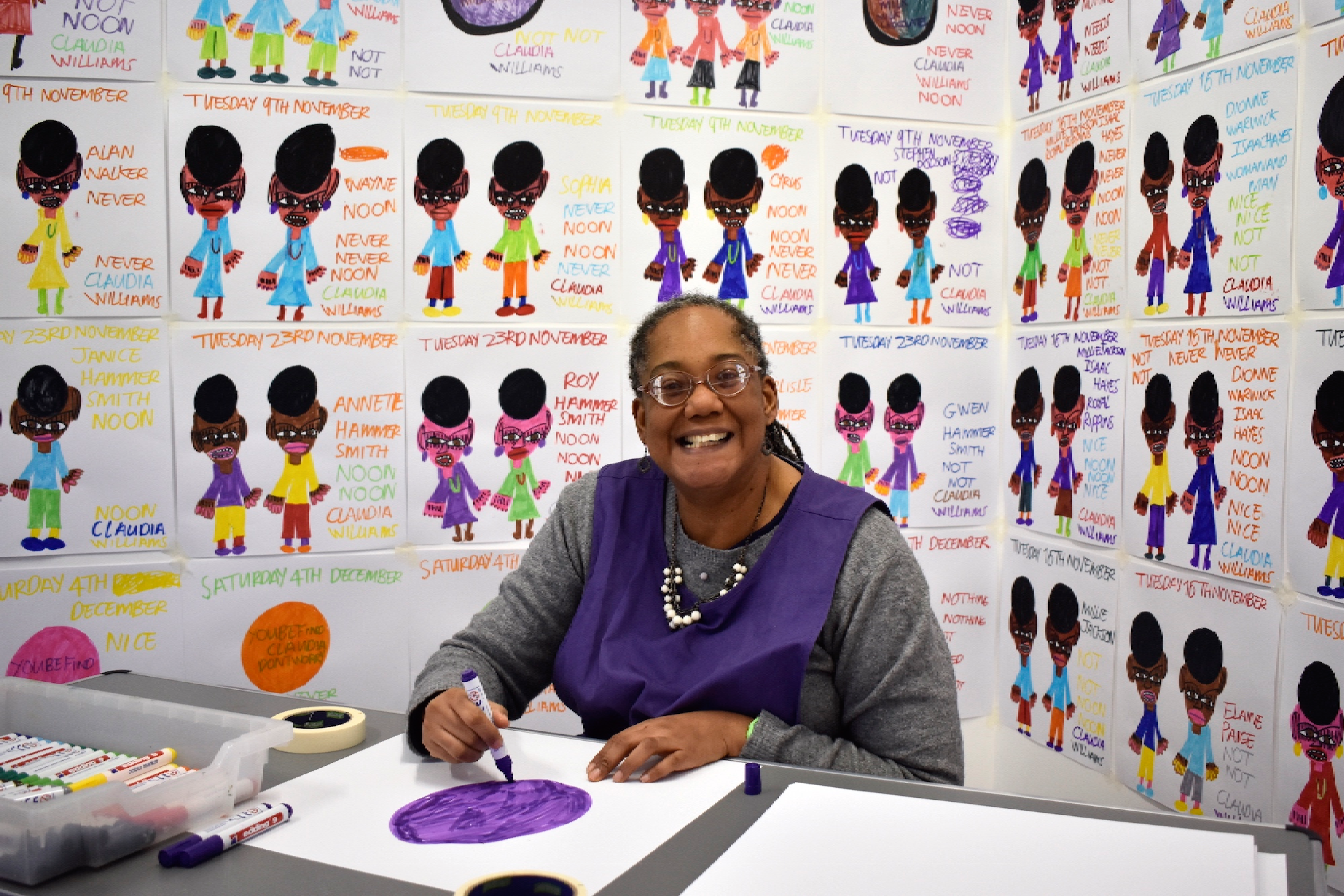 Photograph of Claudia sat at a table drawing. Claudia is looking at the camera smiling. She is surrounded by her drawings which are up on the wall.