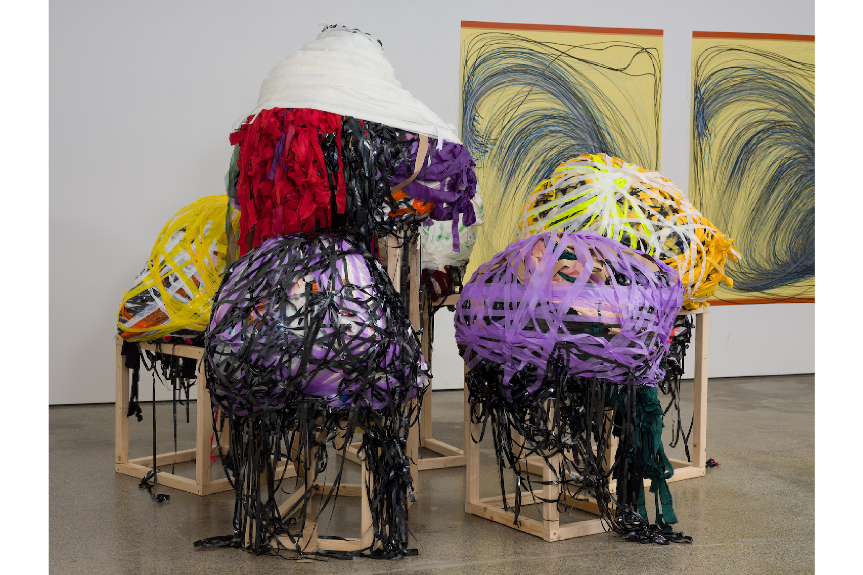 In a gallery, six multimedia wrapped sculptures made with VHS magnetic cassette tape sit on wooden frame plinths.