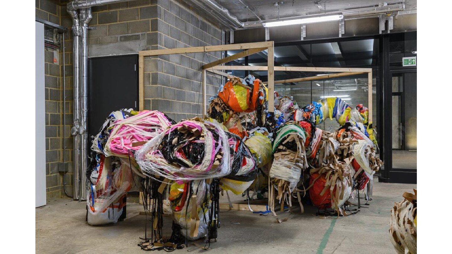 A large sculpture in an industrial gallery space hangs from a wooden structure. Its organic form in made up of various wrapped materials.