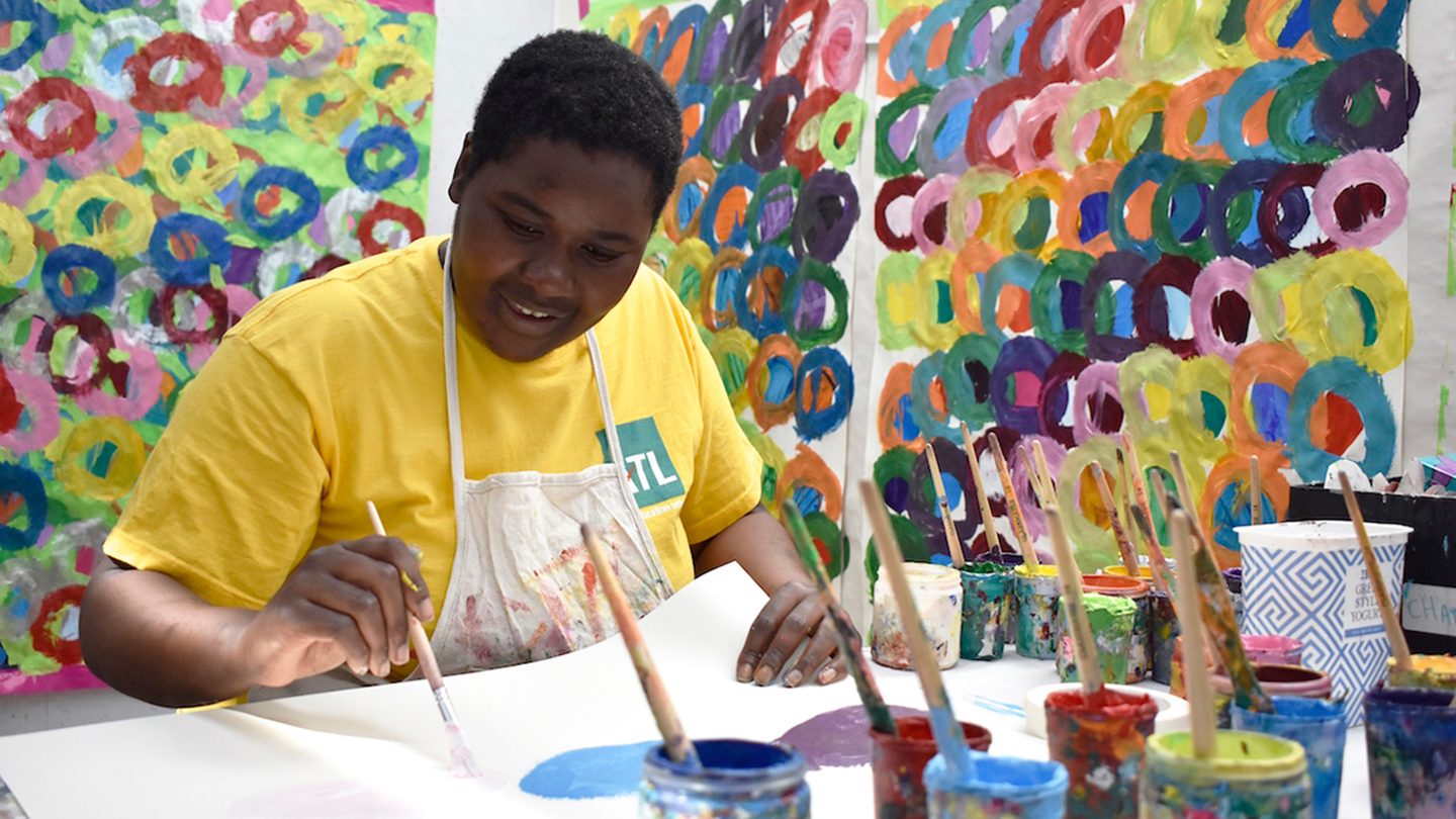 Kwaga sits near a table at Studio Voltaire, working on her art. There are lots of paint pots in front of her, and her colourful artworks are on the wall behind her.