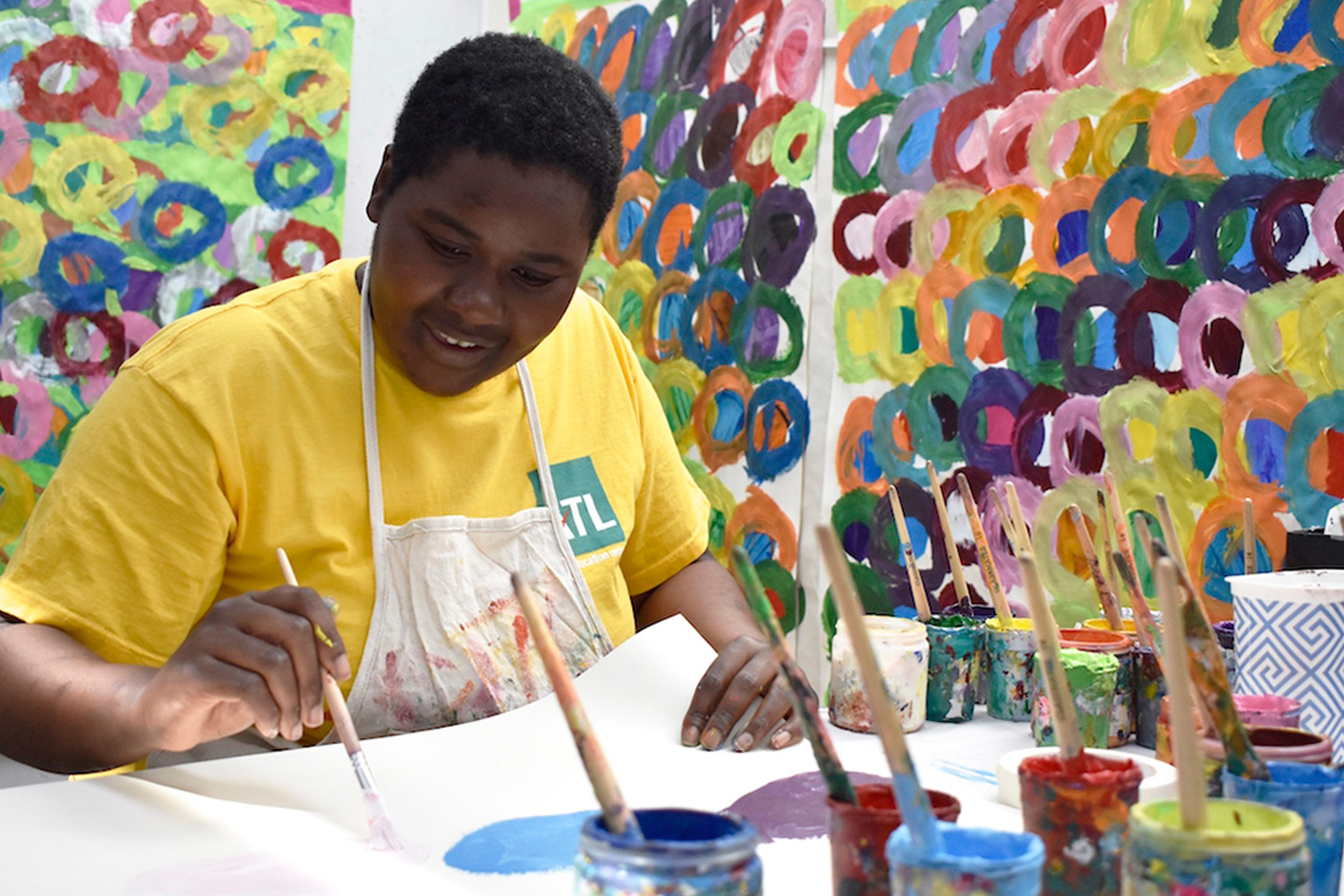 Kwaga sits near a table at Studio Voltaire, working on her art. There are lots of paint pots in front of her, and her colourful artworks are on the wall behind her.