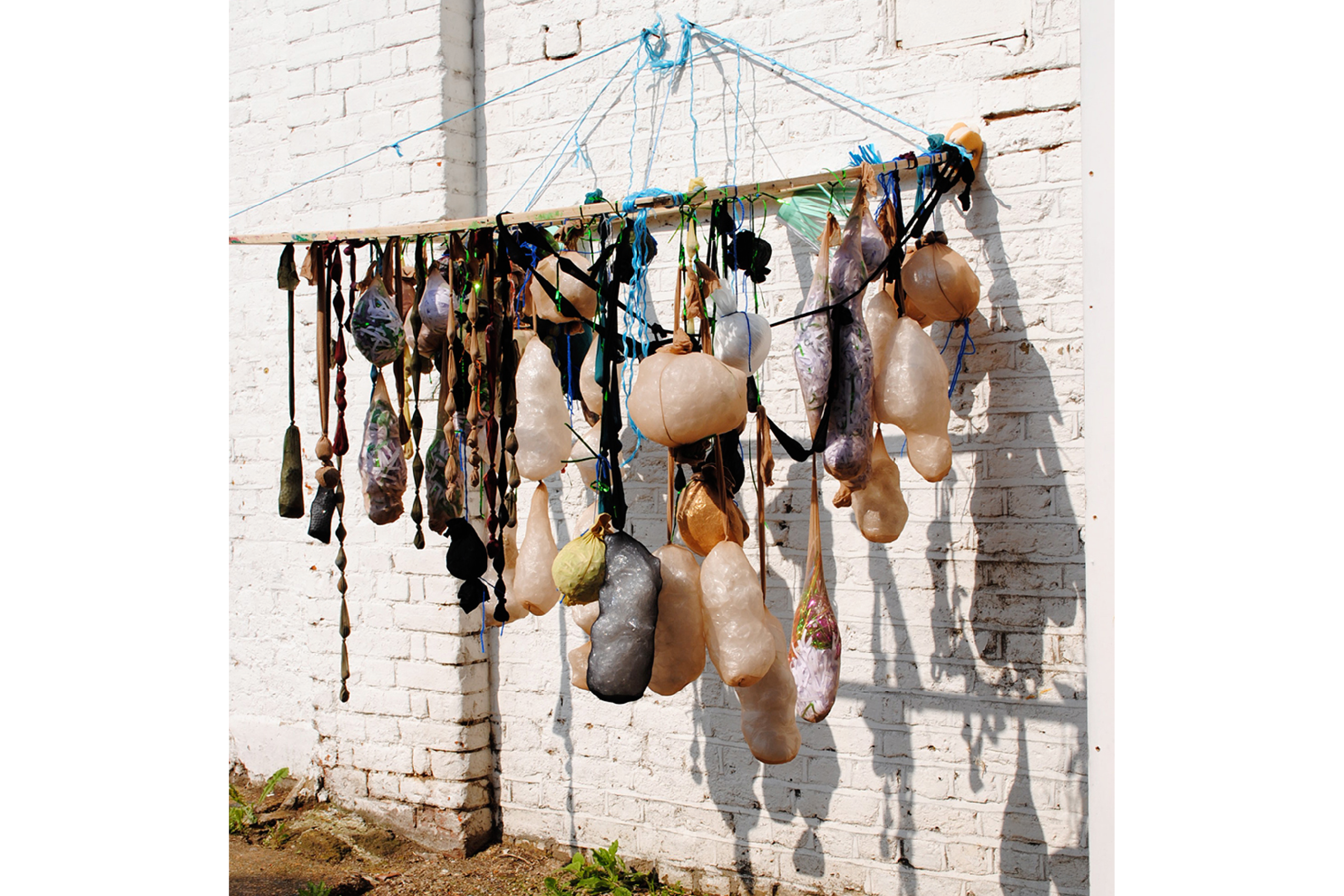 A hanging sculpture against a outdoor white brick wall with strings of stuffed nylon tights suspended from a plank of wood.