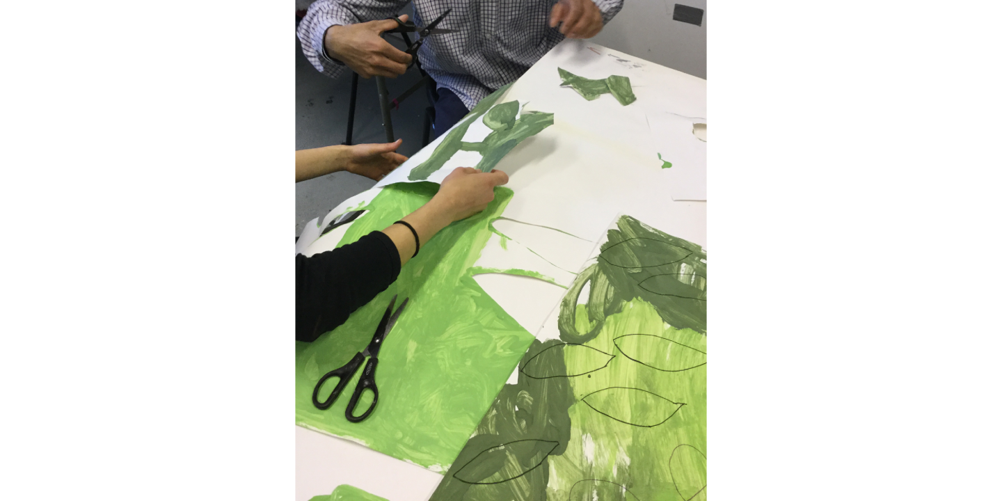 Two people working at a desk, cutting out leaves from paper painted in different hues of green.