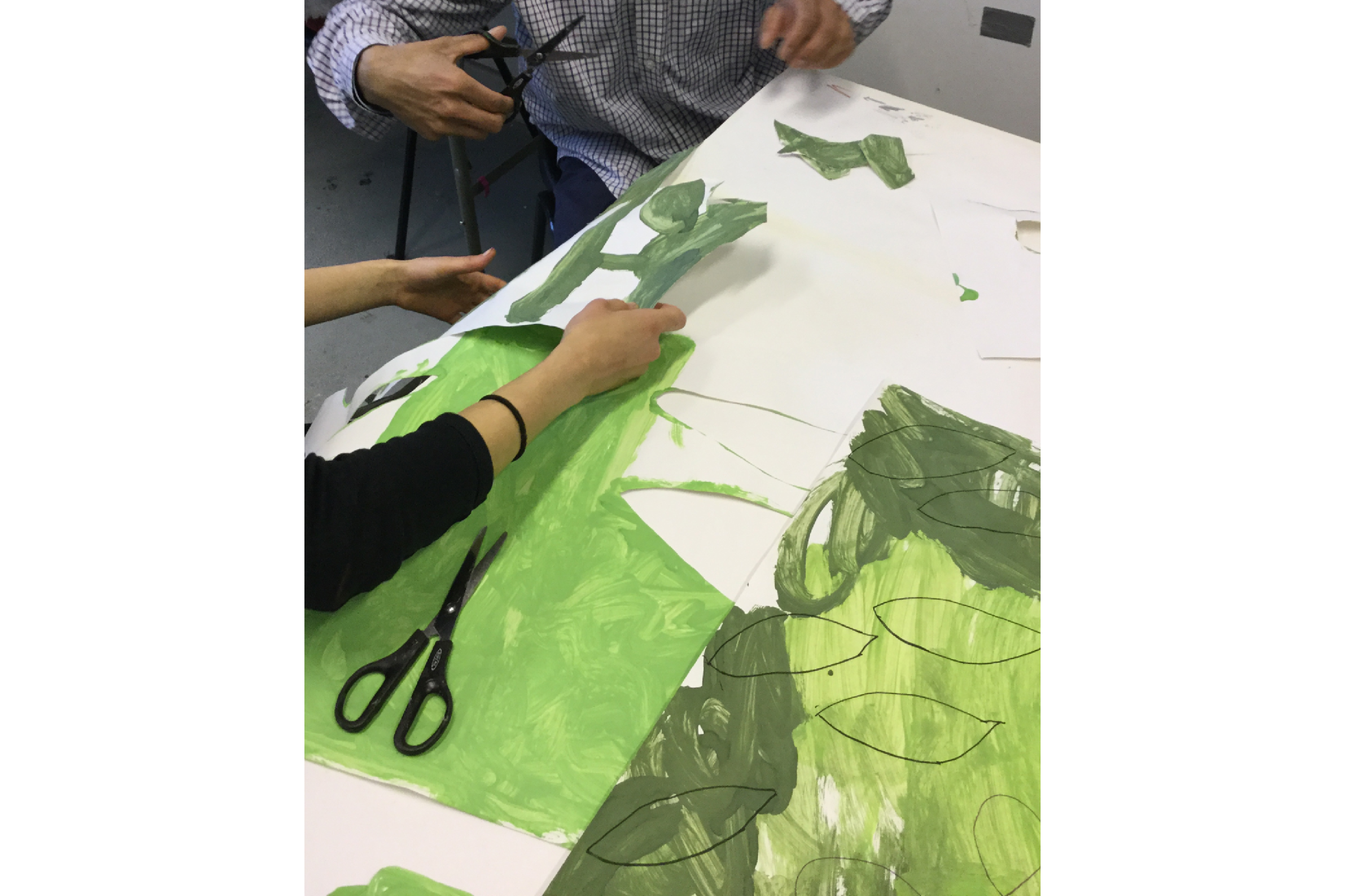 Two people working at a desk, cutting out leaves from paper painted in different hues of green.