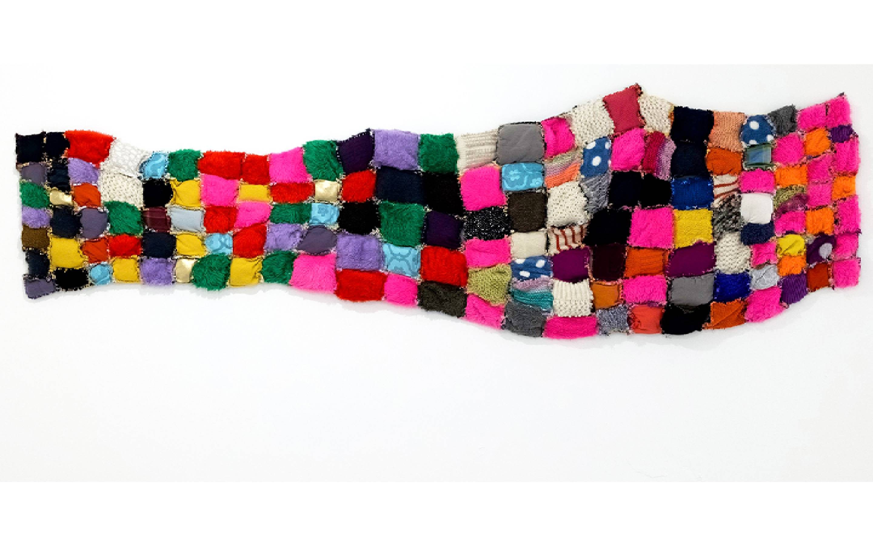 A textile artwork hanging horizontally on a blank white wall. Dozens of colourful textile patches of varying fabrics are sewn together in a soft, grid-like fashion.