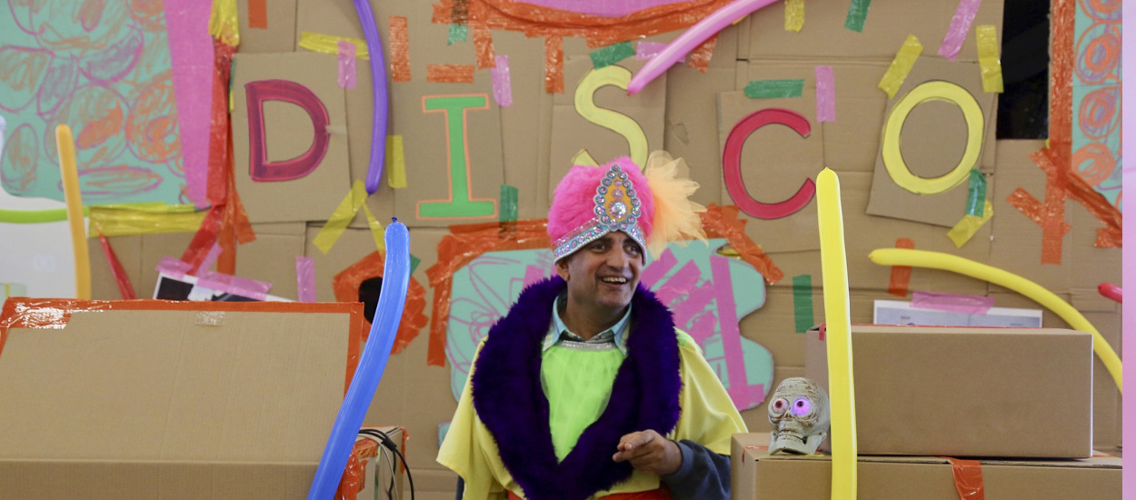 Artist Pardip Kapil wearing a pink turban and yellow smoking jacket with a purple trim in front of a cardboard display that reads "DISCO".