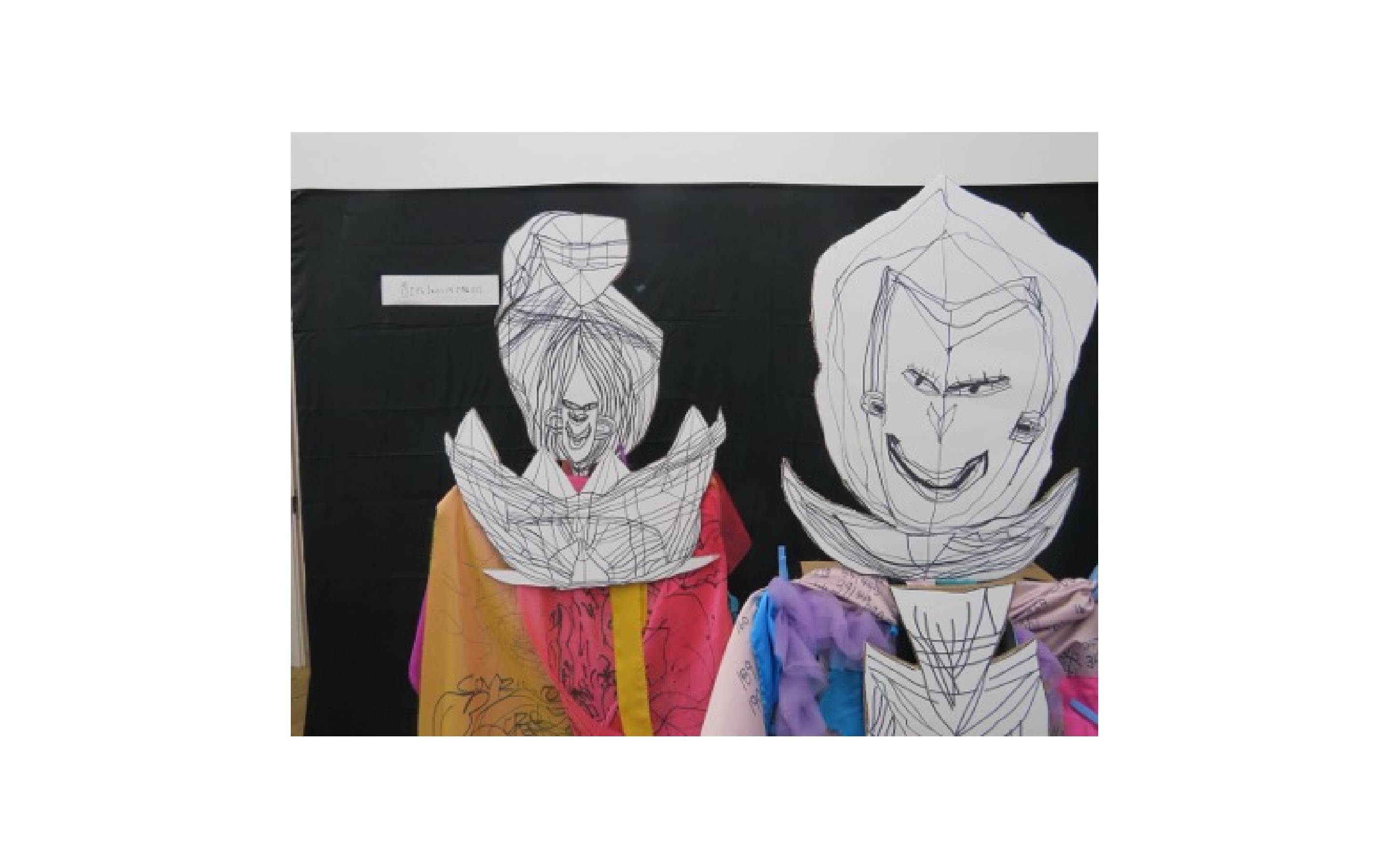 Two people wearing paper masks with black line drawing, whilst draped in colourful fabrics that have pens marks on.