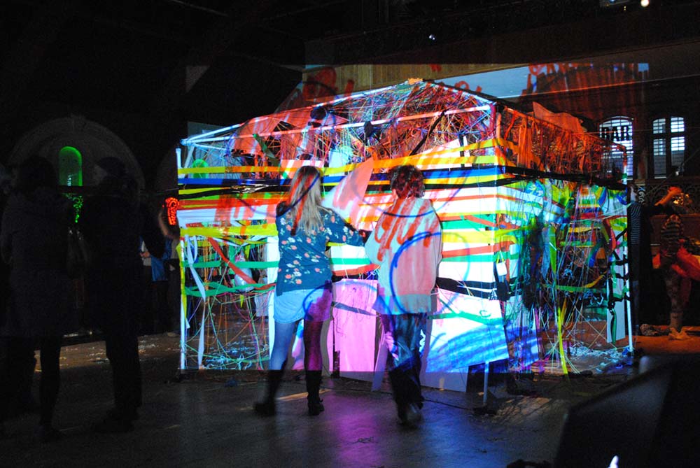 Two people stood in front of an installation in a dark room of a building like structure wrapped in string, coloured tape and paper. that has abstract video projected onto it.