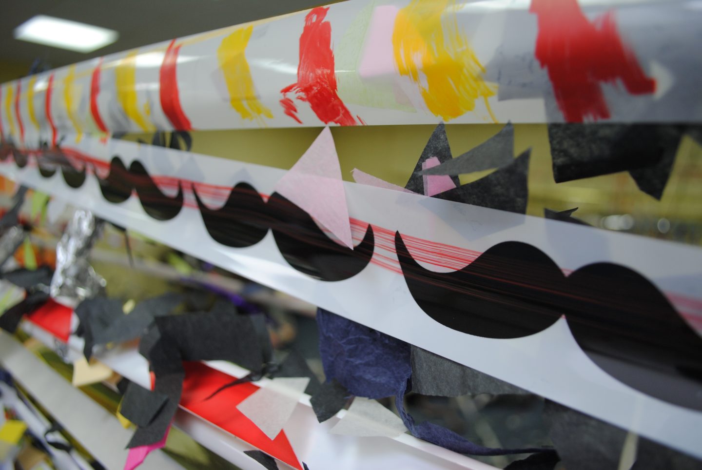 A close up of an artwork made of rows of white tape with dabs of red and yellow paint, cartoon moustaches, and paper applied to it.