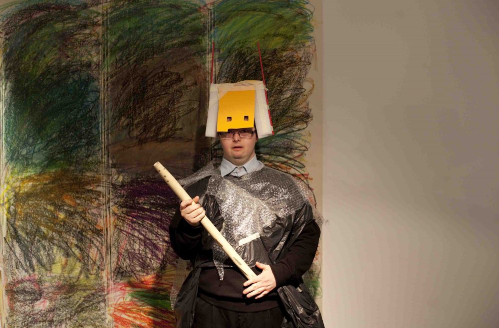 a man stands facing the camera, holding a wooden pole and wearing a colourful cardboard mask.