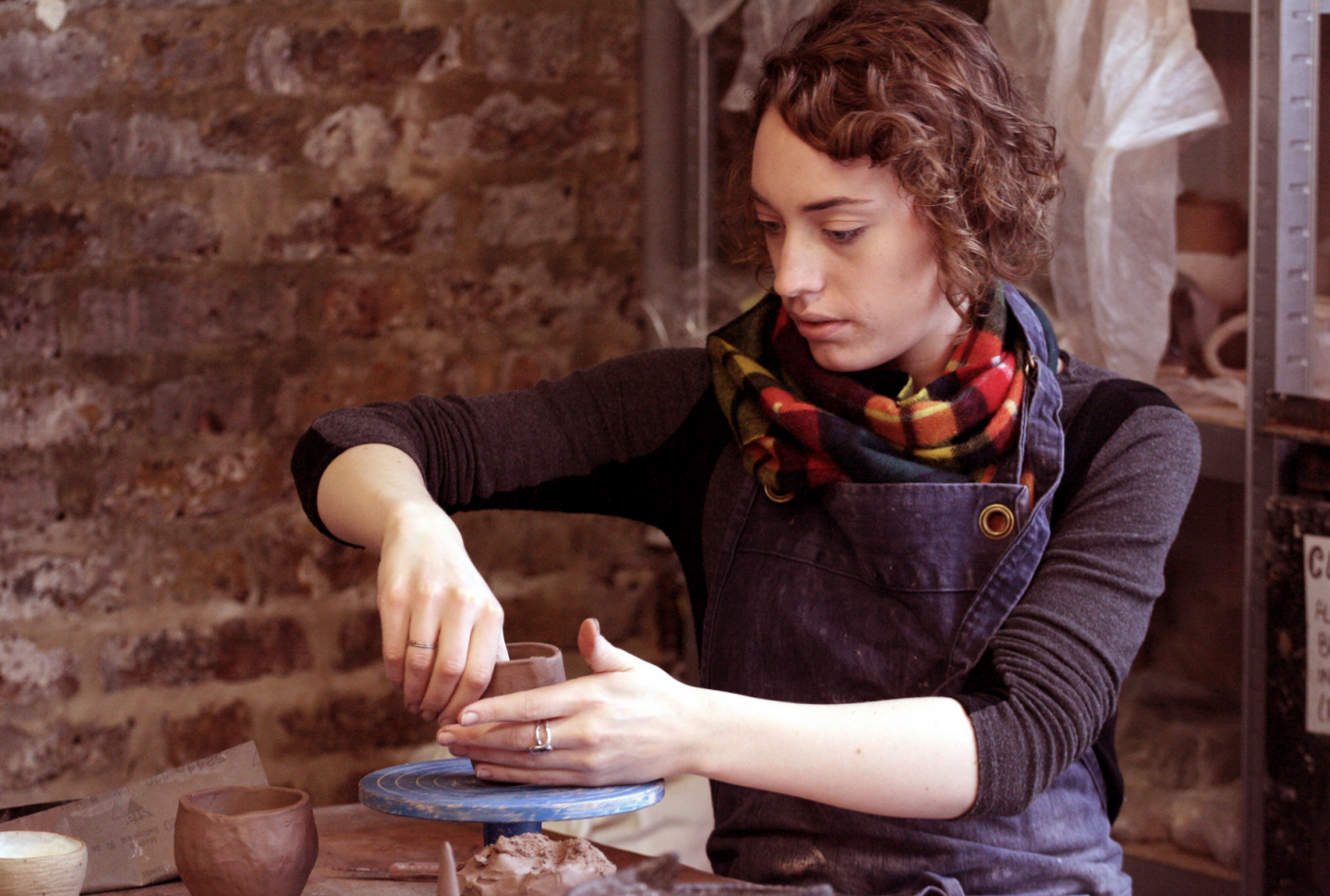 A woman sits near a pottery wheel, making something out of clay